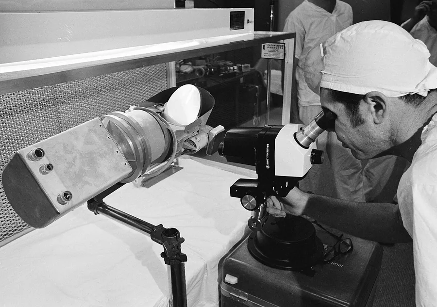 View of scientist looking at a Surveyor III components with a microscope in a Lunar Receiving Laboratory…