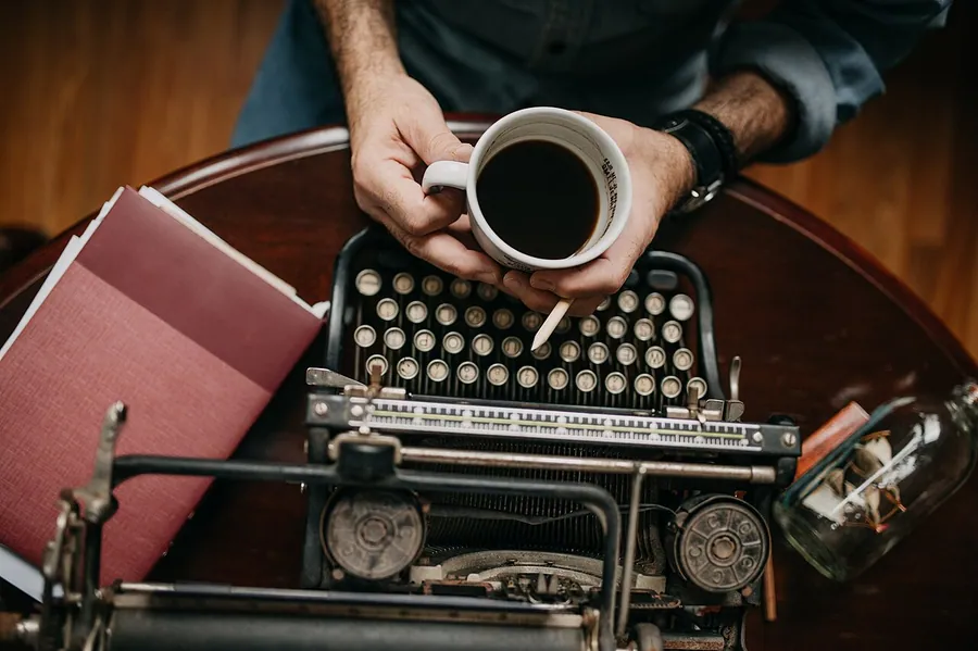 Old man holding cup of coffe near typewriter. Top view.. Фото: Shixart1985 / CC BY 2.0, via Wikimedia Commons