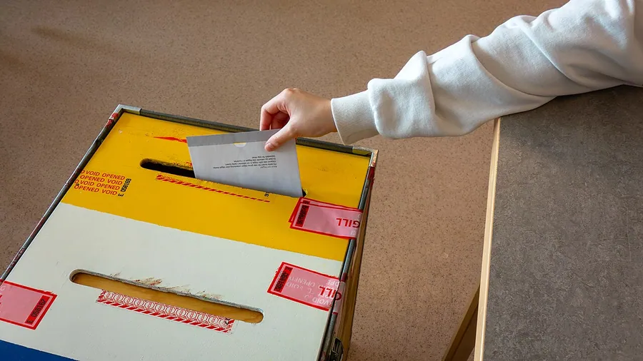 A ballot enclosed in an envelope being dropped into a sealed ballot box by a voting official. The 2024…