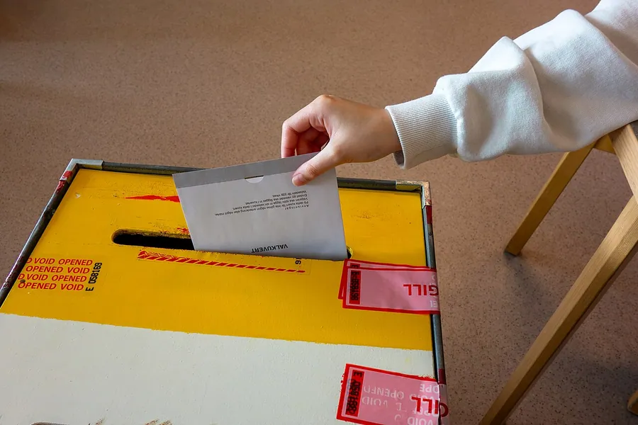 A ballot enclosed in an envelope being dropped into a sealed ballot box by a voting official. The 2024…