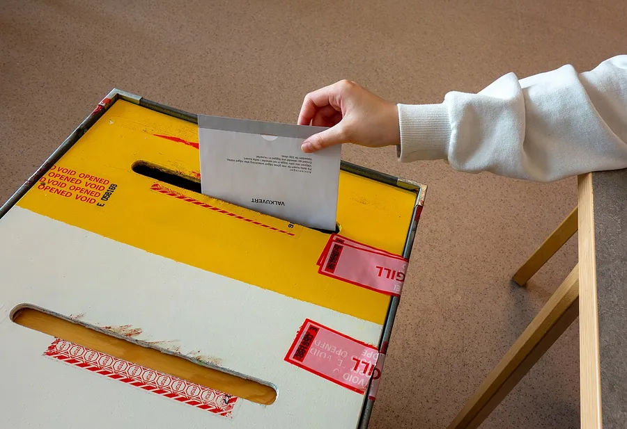 A ballot enclosed in an envelope being dropped into a sealed ballot box by a voting official. The 2024…